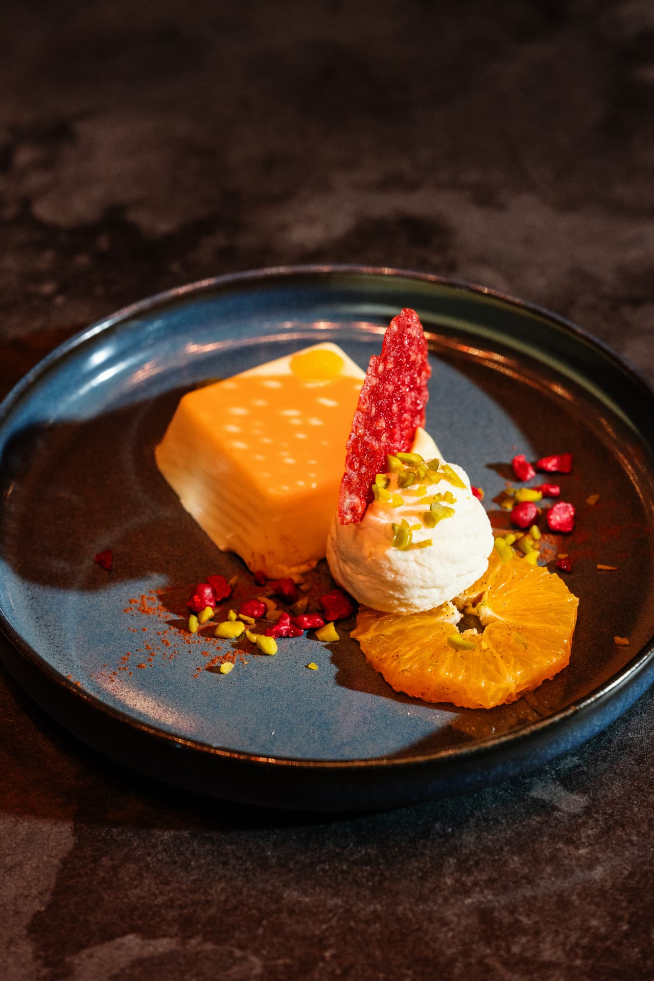 Stilvoll angerichtetes Dessert mit Orangenscheibe und Eiskugel auf einem dunkelblauem Keramikteller.
